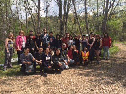 Community Members gathered at our most recent tree planting in a group, in three rows. Behind them, there are large trees without leaves and ahead of the brown earth