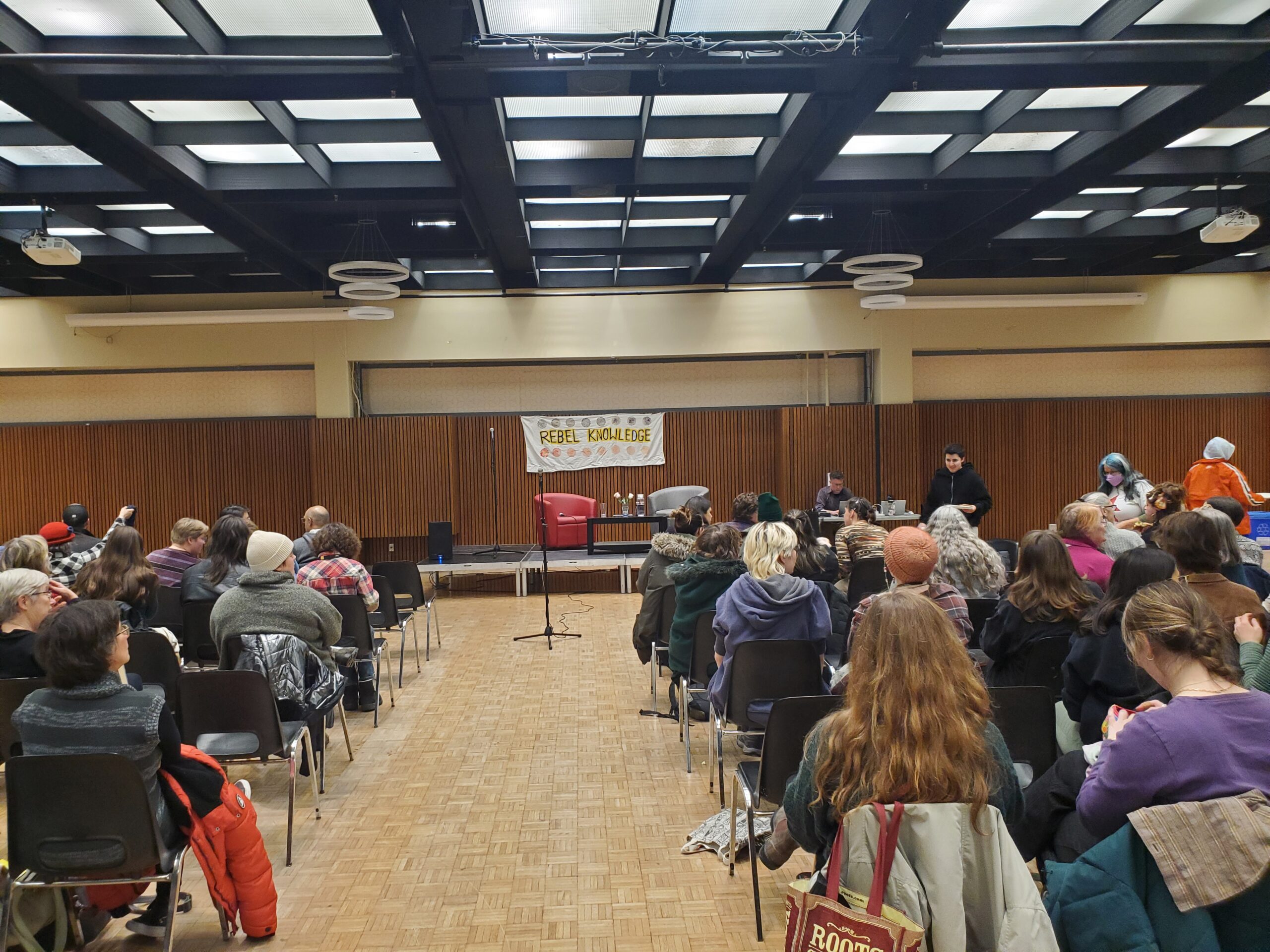 Image shows a large hall filled with people facing a small stage. In the background, there is a banner that reads "Rebel Knowledge" 